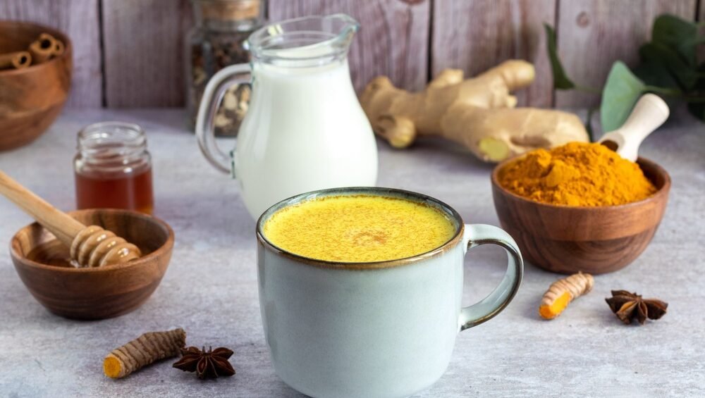 Golden turmeric latte in a light blue mug, surrounded by ingredients including fresh ginger root, honey with honey dipper, turmeric powder in a wooden bowl, milk in a glass pitcher, star anise, and dried turmeric root on a gray concrete surface with rustic wooden background