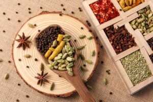 Assorted Indian spices including black peppercorns, turmeric, cardamom pods, and star anise arranged on a wooden spoon placed on a circular wood slice, with a wooden spice box (masala dabba) containing various colorful spices and herbs visible in the corner on a burlap background